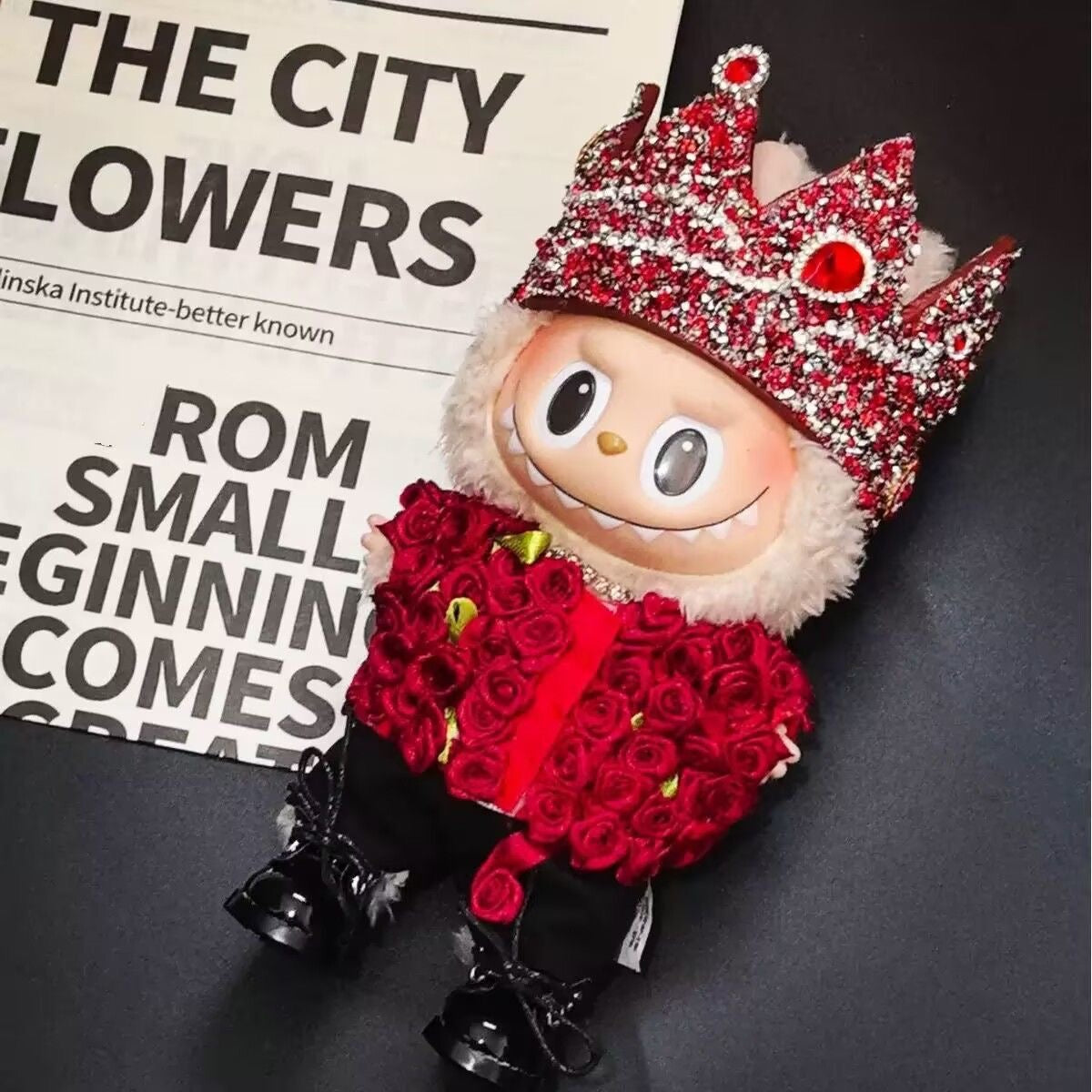 Labubu wearing a handmade G-Dragon inspired outfit – bold K-pop streetwear style with detailed accessories. Red Rose and Crown near newspaper
